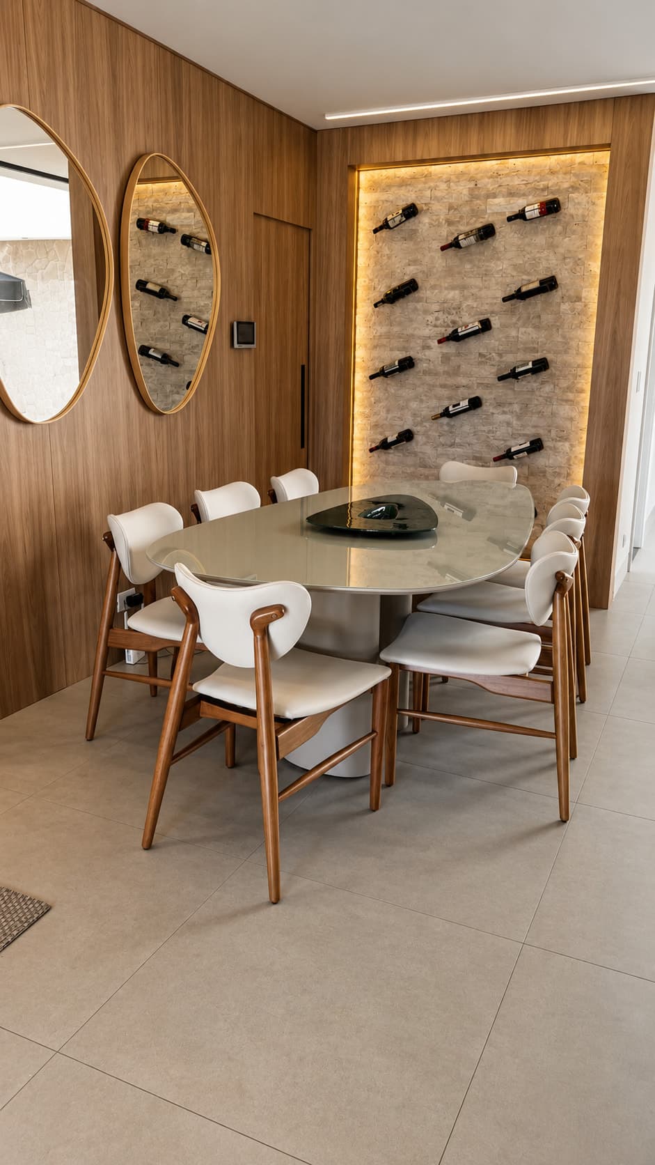 Sala de jantar com mesa oval, painel amadeirado, espelhos redondos e adega de parede iluminada